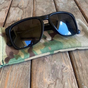 May include: Black sunglasses with a camouflage fabric case. The sunglasses have a rectangular frame and dark lenses.