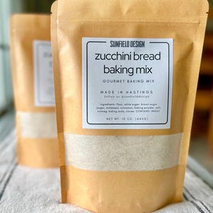 May include: Two brown paper bags of zucchini bread baking mix. The label reads "SUNFIELD DESIGN zucchini bread baking mix" with the net weight of 15 oz. (445g). The ingredients include flour, sugar, cinnamon, and nutmeg.
