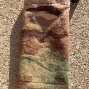 K&ouml;nnte beinhalten: Eine Camouflage-Kordelzugtasche mit einem schwarzen Kordelzug und einem Knopf. Die Tasche ist aus einem weichen, strapazierf&auml;higen Stoff.