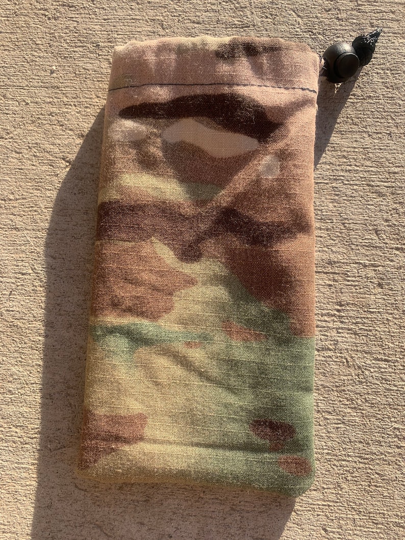 K&ouml;nnte beinhalten: Eine Camouflage-Kordelzugtasche mit einem schwarzen Kordelzug und einem Knopf. Die Tasche ist aus einem weichen, strapazierf&auml;higen Stoff.