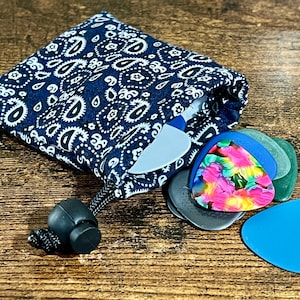 Puede incluir: Una bolsa con cordón de color azul marino con estampado de cachemira y una colección de púas de guitarra. Las púas son de varios colores, incluyendo azul, verde y un diseño multicolor vibrante. La bolsa está abierta.