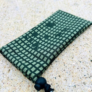 May include: A green drawstring pouch with a geometric pattern and a black drawstring. The pouch is folded and lying on a light brown surface.