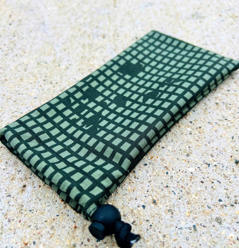 May include: A green drawstring pouch with a geometric pattern and a black drawstring. The pouch is folded and lying on a light brown surface.