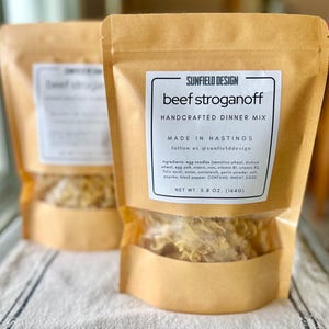 Könnte beinhalten: Zwei Kraftpapierbeutel mit handgemachter Beef Stroganoff-Dinner-Mischung. Jeder Beutel hat unten ein klares Fenster, das den Inhalt zeigt. Das Etikett lautet "SUNFIELD DESIGN beef stroganoff HANDCRAFTED DINNER MIX". Nettogewicht 164 Gramm.