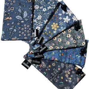 May include: A collection of drawstring bags with floral patterns in various shades of blue, gray, and yellow. Each bag features a black drawstring closure and a small tag with the text "SUNFIELD DESIGN". The bags are arranged in a fan shape.