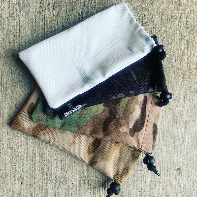 K&ouml;nnte beinhalten: Vier verschiedene Stoffbeutel, einer wei&szlig;, einer schwarz, einer Tarnmuster und einer beige. Der Tarnmusterbeutel hat ein schwarzes Etikett mit dem Text "SD SUNFIELD DESIGN".