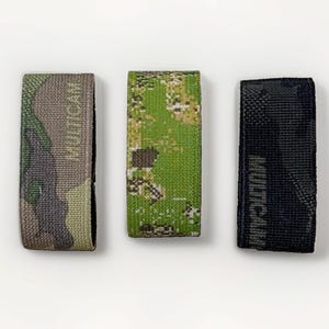 May include: Three rectangular fabric straps featuring camouflage patterns. The left strap has a tan and green camo print with the text MULTICAM. The center strap has a bright green and brown camo print. The right strap is black with the text MULTICAM.