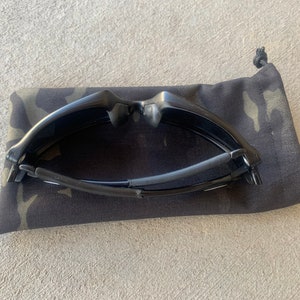 K&ouml;nnte beinhalten: Schwarze Sonnenbrille, zusammengeklappt und in einem schwarzen und gr&uuml;nen Tarnmuster-Etui.