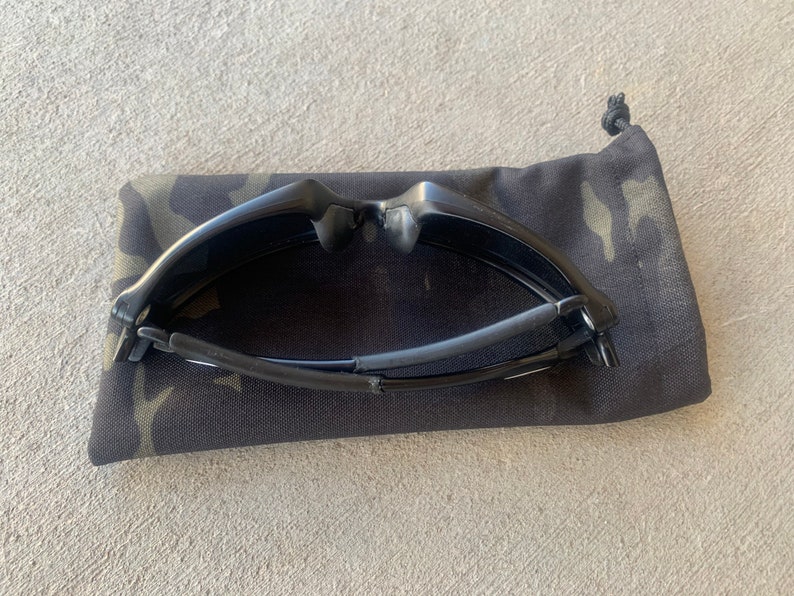 K&ouml;nnte beinhalten: Schwarze Sonnenbrille, zusammengeklappt und in einem schwarzen und gr&uuml;nen Tarnmuster-Etui.