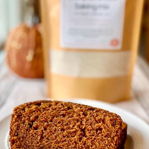 Puede incluir: Una rebanada de pan de calabaza en un plato blanco. El pan es de color marrón cálido con una superficie texturizada. En el fondo, se ve una bolsa de mezcla para hornear con especias de calabaza, junto con una calabaza decorativa.