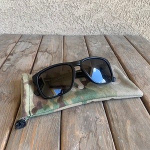 K&ouml;nnte beinhalten: Schwarze Sonnenbrille mit einem Etui aus Tarnstoff. Die Sonnenbrille hat dunkelgraue Gl&auml;ser und einen schwarzen Rahmen.