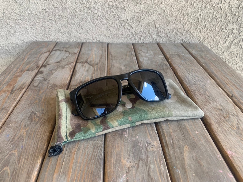 K&ouml;nnte beinhalten: Schwarze Sonnenbrille mit einem Etui aus Tarnstoff. Die Sonnenbrille hat dunkelgraue Gl&auml;ser und einen schwarzen Rahmen.