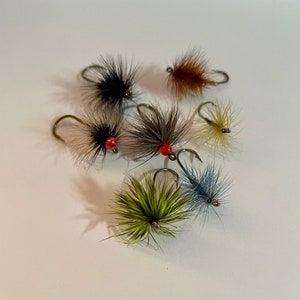 May include: Seven artificial fishing flies in a variety of colours. The flies have fluffy, textured bodies in black, brown, green, and blue, with curved metal hooks. Some flies have red beads.