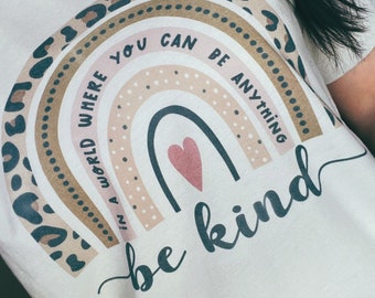 May include: A white t-shirt featuring a rainbow design and the text "In a world where you can be anything, be kind". The rainbow is made up of brown, pink, and white stripes with a leopard print border. A small pink heart is in the centre of the rainbow.