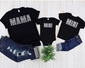 May include: Black t-shirts with white "MAMA" and "MINI" text in a leopard print design. The shirts are on a white wooden surface with a small plant in the middle.