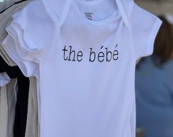 May include: White baby onesie with black text that reads "the bébé".