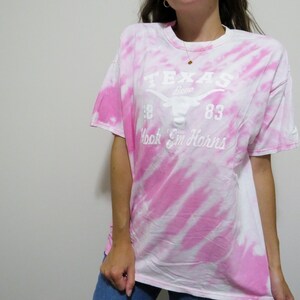 Puede incluir: Una camiseta te&ntilde;ida con tinte blanco y rosa con el texto "TEXAS" encima de un logotipo de cuerno largo y las palabras "Hook 'Em Horns". La camiseta tiene mangas cortas y cuello redondo.