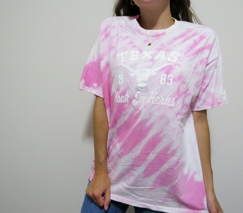 Puede incluir: Una camiseta te&ntilde;ida con tinte blanco y rosa con el texto "TEXAS" encima de un logotipo de cuerno largo y las palabras "Hook 'Em Horns". La camiseta tiene mangas cortas y cuello redondo.
