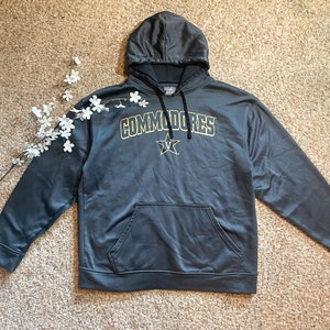 May include: A dark gray hooded sweatshirt with the word "COMMODORES" in gold lettering and a gold star logo. The sweatshirt has a front pocket and a drawstring hood. A sprig of white flowers is placed on the left side of the sweatshirt.