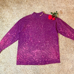 May include: A long-sleeved purple shirt with a mock neck and a speckled pink pattern. The word "GREATER" is embroidered in gold thread on the collar. A few red roses are placed on the shirt.