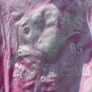Puede incluir: Camiseta rosa te&ntilde;ida con letras blancas y un logotipo de Longhorn. El texto incluye "TEXAS", "83" y "Hook 'Em Horns". La camiseta tiene un aspecto descolorido y vintage.