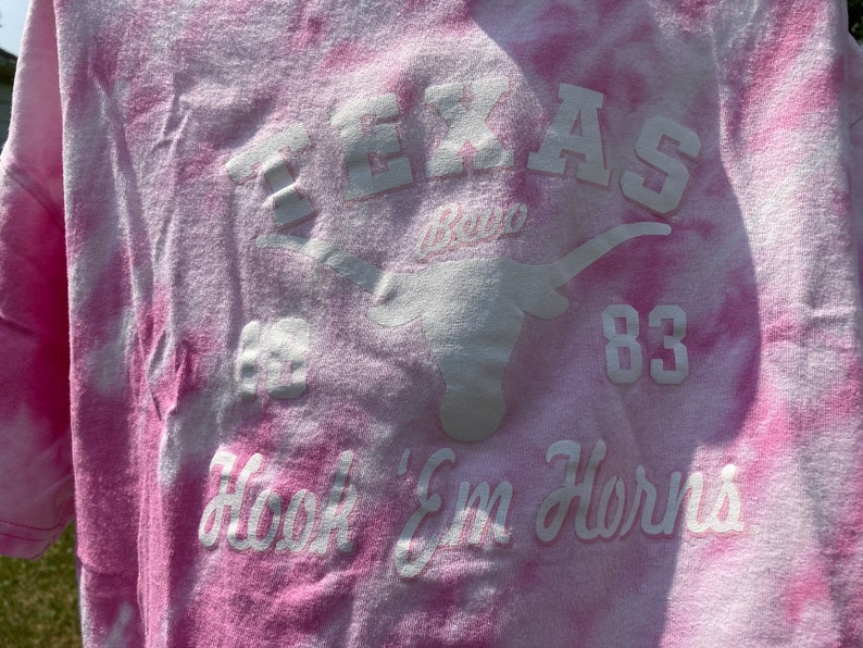 Puede incluir: Camiseta rosa te&ntilde;ida con letras blancas y un logotipo de Longhorn. El texto incluye "TEXAS", "83" y "Hook 'Em Horns". La camiseta tiene un aspecto descolorido y vintage.