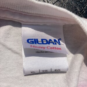 Puede incluir: Primer plano de una etiqueta de camiseta blanca con la marca "GILDAN" en azul. La etiqueta dice "Heavy Cotton" y "Hecho en Honduras". Se muestran las tallas XL, TG y EG. La camiseta tiene un estampado tie-dye.