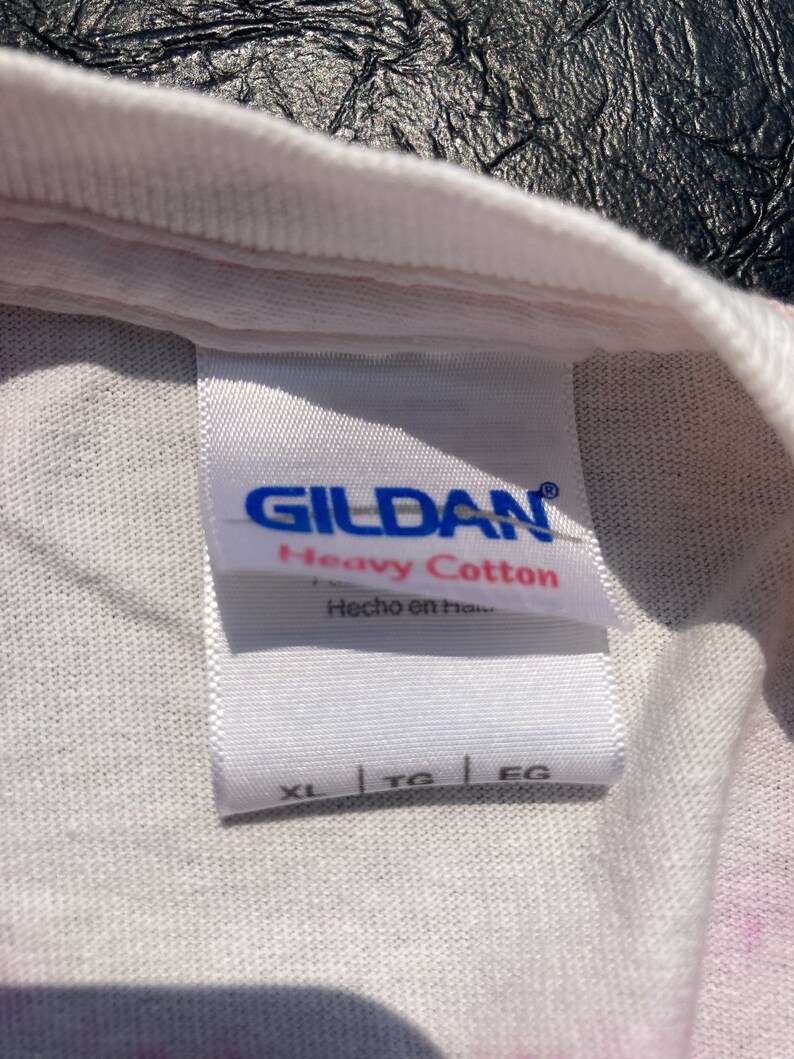 Puede incluir: Primer plano de una etiqueta de camiseta blanca con la marca "GILDAN" en azul. La etiqueta dice "Heavy Cotton" y "Hecho en Honduras". Se muestran las tallas XL, TG y EG. La camiseta tiene un estampado tie-dye.