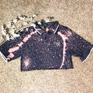 May include: A navy blue cropped polo shirt with a bleach-splatter design and white trim. The shirt features a small logo on the chest and a white collar. The short sleeves have white trim. The shirt is a unique, casual style.
