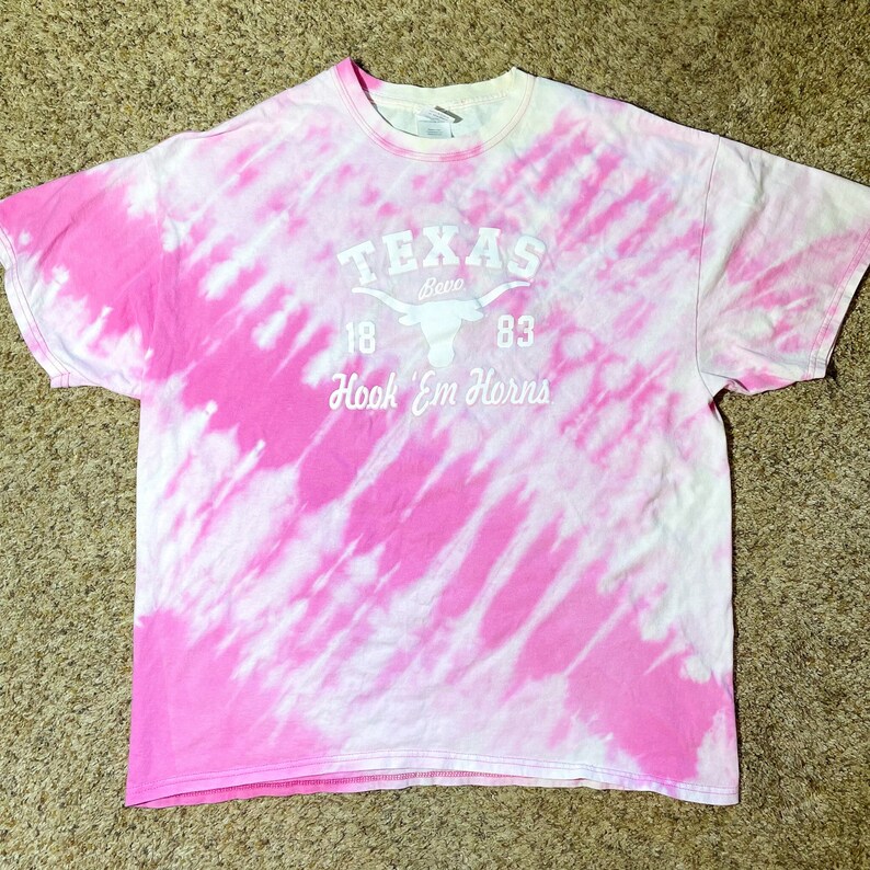 Puede incluir: Una camiseta te&ntilde;ida con tinte rosa y blanco con el texto "TEXAS" encima de un gr&aacute;fico de cuerno largo y las palabras "Bevo 18 83 Hook 'Em Horns" en blanco. La camiseta es de manga corta y tiene cuello redondo.