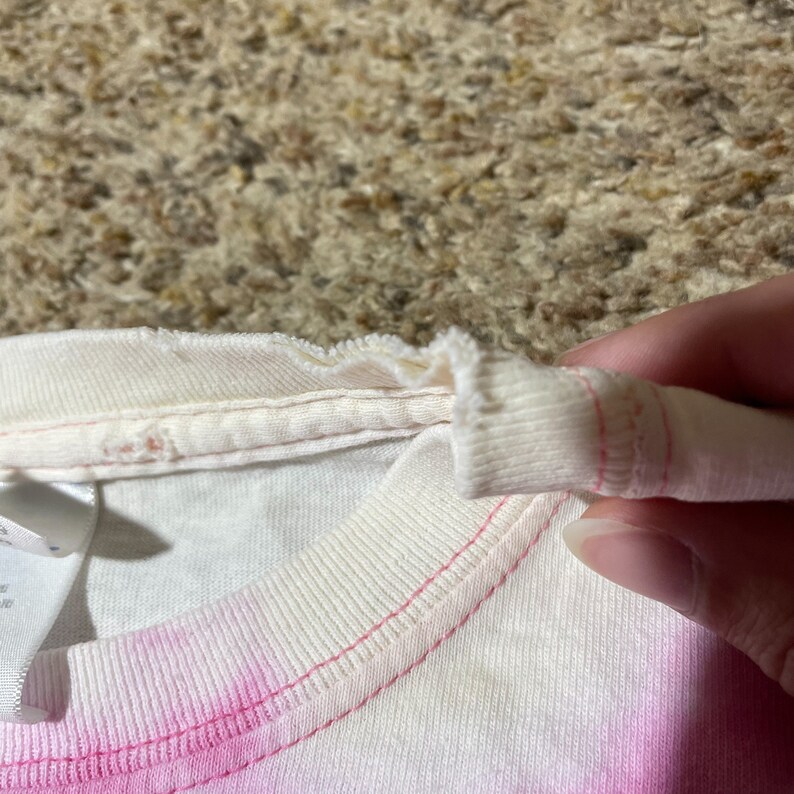 Puede incluir: Primer plano del cuello de una camiseta te&ntilde;ida con tinte blanco y rosa. El cuello es acanalado y tiene costuras rosas. La camisa est&aacute; sujeta por una mano.