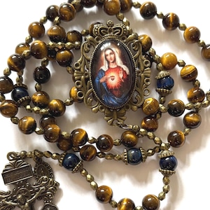 Op de afbeelding: Een rozenkrans met tijgeroogkralen en een gedetailleerd kruisbeeld. Het middelpunt toont een afbeelding van de Maagd Maria. De kralen zijn een mix van bruin en goud, met enkele donkerblauwe kralen.