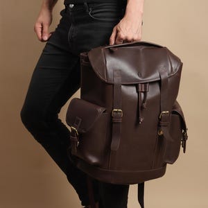 May include: A brown leather backpack with multiple compartments and adjustable straps. The backpack features a flap closure with a buckle and a drawstring closure. The bag has two side pockets and a top handle. The hardware is gold-colored.