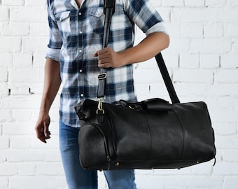 May include: A black leather duffel bag featuring a long adjustable shoulder strap with a padded section, gold-toned metal hardware, side zipped pockets, and protective metal feet on the bottom base for durability and structure.