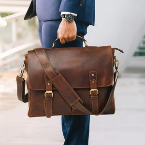 May include: A brown leather briefcase with a shoulder strap and gold-colored hardware. The briefcase has a flap closure with two buckle straps. The bag is held by a person wearing a blue suit.