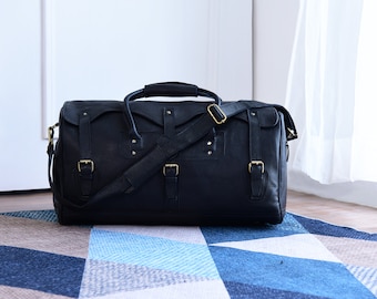 May include: A black leather duffel bag with gold-coloured buckles and hardware. The bag has a top handle and a detachable shoulder strap. The bag is rectangular and appears to be made of high-quality leather. The bag is on a blue and white patterned rug.