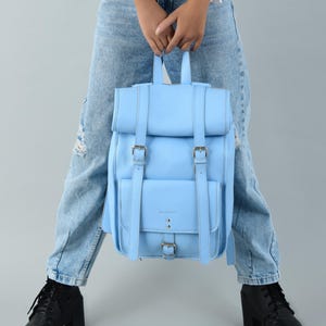 May include: A light blue leather backpack with a flap closure, two front pockets, and adjustable straps. The backpack has silver buckles and is held by a person wearing light blue jeans and black boots.