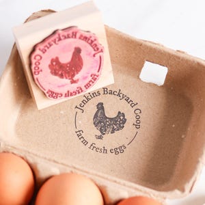 May include: A wooden rubber stamp with a circular design featuring a chicken and the text Jenkins Backyard Coop farm fresh eggs, positioned above a matching brown paper egg carton stamped with the same black ink design and brown eggs.