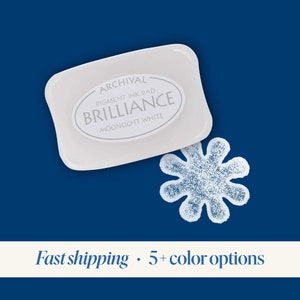 K&ouml;nnte beinhalten: Archival Pigment Tintenkissen in Moonlight White von Brilliance. Das Tintenkissen ist wei&szlig; und hat einen blauen Hintergrund. Das Tintenkissen hat die Form eines Rechtecks mit abgerundeten Ecken. Das Tintenkissen liegt auf einer blauen Oberfl&auml;che.