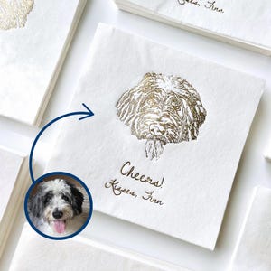 May include: White paper napkin with a gold foil illustration of a dog's head. The text "Cheers! Kisses, Finn" is written in gold foil below the dog.