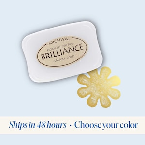 K&ouml;nnte beinhalten: Ein wei&szlig;es Stempelkissen mit dem Text "ARCHIVAL PIGMENT INK PAD BRILLIANCE GALAXY GOLD" darauf. Das Stempelkissen liegt neben einem goldenen sternf&ouml;rmigen Stempel.