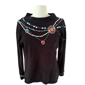 Könnte beinhalten: Schwarzer Pullover mit Stehkragen, verziert mit bunten Perlenverzierungen. Das Design umfasst eine zentrale kreisförmige Brosche und kaskadierende Perlenstränge mit roten, blauen und weißen Akzenten. Die Manschetten haben ebenfalls Perlendetails.