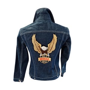 May include: Dark blue denim jacket with white stitching. The back features a large embroidered patch of a golden eagle with the words "GOLD HONDA WING" in red and white. The jacket has a classic collar and button closures.