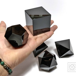 Puede incluir: Cuatro formas de cristal de obsidiana negra, incluyendo un cubo, un dodecaedro y dos pirámides. Los cristales están sobre una superficie blanca.
