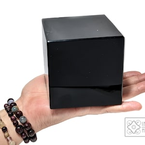May include: A large, black obsidian cube, approximately 3 inches in size, is held in a person's hand. The cube is smooth and polished, with a reflective surface.