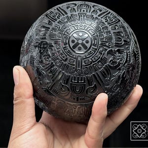 Könnte beinhalten: Schwarze Keramikskugel mit aufwendigen Schnitzereien, die ein kreisförmiges Design mit einem zentralen Symbol darstellen. Die Kugel wird in einer Hand gehalten.