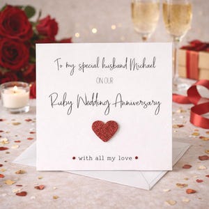 May include: A white square greeting card with black text reading To my special husband Michael ON OUR Ruby Wedding Anniversary and with all my love. The card features a central red glitter heart and two small red dots on the bottom line.