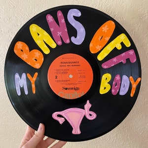 May include: A black vinyl record with the words "Bans Off My Body" painted in colorful letters with flowers. The record also features a pink illustration of a uterus.