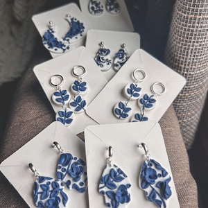 May include: A collection of handmade earrings with white and blue floral designs. The earrings are displayed on white cards, showcasing various shapes and styles, including teardrops and circular designs. The earrings have silver-colored hooks.