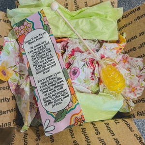 Op de afbeelding: Een roze en witte inspirerende bladwijzer met een bloemenpatroon en de tekst "Richt je ogen op Jezus" en een citaat over het vinden van licht in de duisternis. De bladwijzer zit in een bruine kartonnen doos met tissuepapier en een gele lolly.